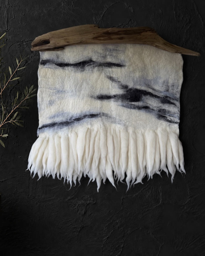 Ivory felt and wool wall hanging with charcoal-gray abstract lines and soft fringe, suspended from natural driftwood on a dark textured wall.