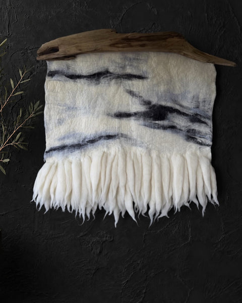Ivory felt and wool wall hanging with charcoal-gray abstract lines and soft fringe, suspended from natural driftwood on a dark textured wall.